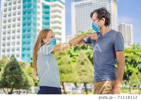 Elbow greeting to avoid the spread of coronavirus COVID-19. A man and a woman in medical face masks meet on the street with bare hands. Instead of greeting with a hug or handshake, they bump elbows Elbow greeting to avoid the spread of coronavirus COVID-19. A man and a woman in medical face masks meet on the street with bare hands. Instead of greeting with a hug or handshake, they bump elbows 71118112