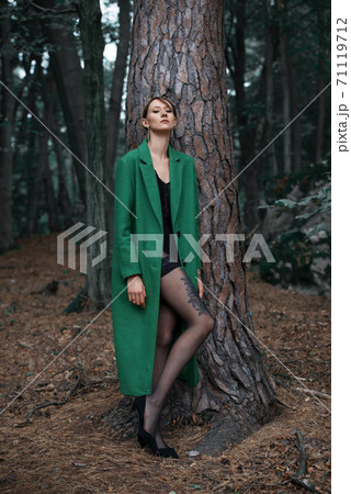 Fashionable young woman in green coat posing in a forest near the tree 71119712