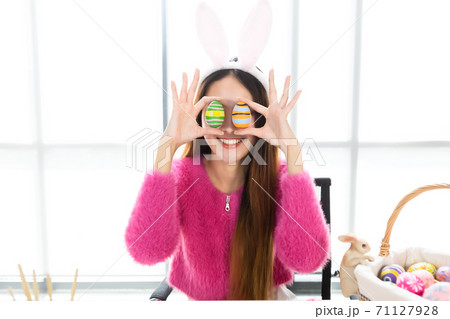 Happy smile Asian Young woman wearing bunny ears and holding up a colorful Easter egg in front of her eye Skewers In the white room background Happy smile Asian Young woman wearing bunny ears and holding up a colorful Easter egg in front of her eye Skewers In the white room background 71127928