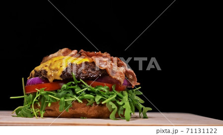 Craft burger is cooking on black background. Consist: sauce, arugula, tomato, onion, bacon, currant sauce, ricotta cheese, air bun and marble meat. Craft burger is cooking on black background. Consist: sauce, arugula, tomato, onion, bacon, currant sauce, ricotta cheese, air bun and marble meat. 71131122