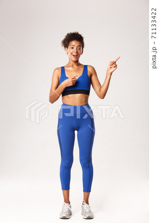 Full length of excited good-looking african-american sportswoman in fitness clothing, pointing fingers at upper right corner, standing over white background Full length of excited good-looking african-american sportswoman in fitness clothing, pointing fingers at upper right corner, standing over white background 71133422