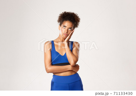 Studio shot of bored or annoyed african-american fitness girl roll eyes up and leaning head on hand, standing unamused against white background 71133430