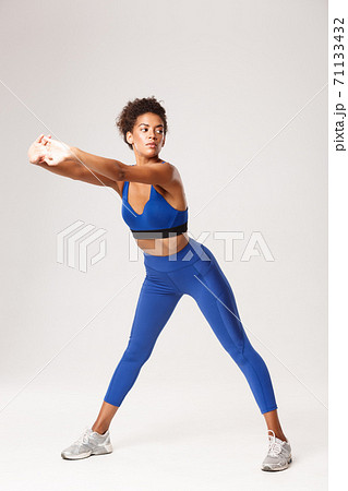 Full length of attractive african-american woman with fit body, wearing blue sportwear, exercising over white background, showing stretching warm-up workout, looking aside 71133432