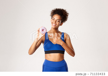 Young smiling fitness woman in blue sport outfit, pointing finger at donut, showing sweets, standing against white background 71133433