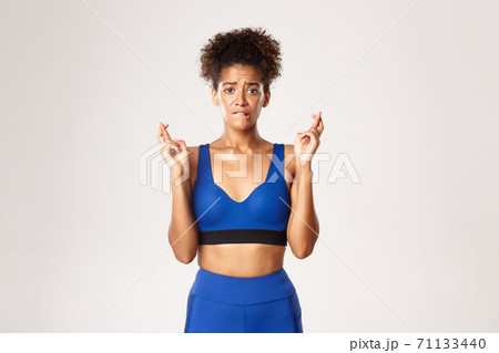 Concept of sport and workout. Hopeful and worried african-american sportswoman, wearing blue sportswear, biting lip and crossing finger for good luck, making wish against white background 71133440