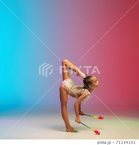 Little caucasian girl, rhytmic gymnast training, performing isolated on gradient blue-red studio background in neon Little caucasian girl, rhytmic gymnast training, performing isolated on gradient blue-red studio background in neon 71134131