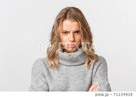 Close-up of angry blond girl in grey sweater, looking from under forehead and sulking, feeling offended, standing over white background Close-up of angry blond girl in grey sweater, looking from under forehead and sulking, feeling offended, standing over white background 71134916