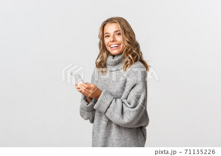 Portrait of attractive happy woman in grey sweater, holding smartphone and smiling at camera, concept of online shopping and social networks Portrait of attractive happy woman in grey sweater, holding smartphone and smiling at camera, concept of online shopping and social networks 71135226