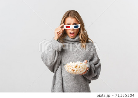 Image of blond girl in 3d glasses, holding bowl with popcorn, watching movie and looking confused, standing over white background Image of blond girl in 3d glasses, holding bowl with popcorn, watching movie and looking confused, standing over white background 71135304
