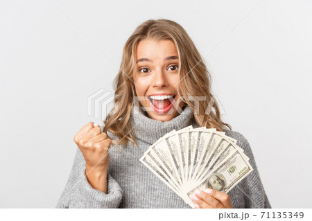 Close-up of successful and rich blond girl in grey sweater, holding money and looking like winner, standing over white background Close-up of successful and rich blond girl in grey sweater, holding money and looking like winner, standing over white background 71135349