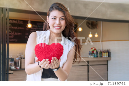 smiling beautiful young woman standing and holding pillow small red heart pillow in front of cafe. 71139741