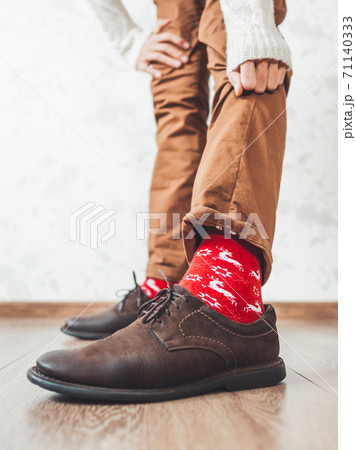 Young man pulls up leg of his chinos trousers to show bright red socks with reindeers on them. Scandinavian pattern. Winter holiday spirit. Casual outfit for New Year and Christmas celebration. Young man pulls up leg of his chinos trousers to show bright red socks with reindeers on them. Scandinavian pattern. Winter holiday spirit. Casual outfit for New Year and Christmas celebration. 71140333