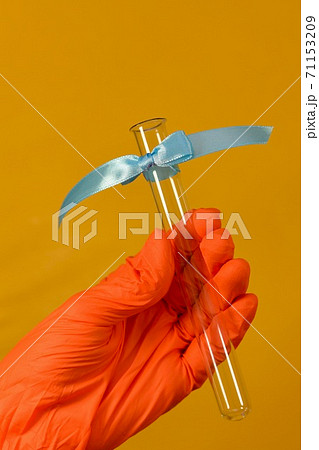 A hand in an orange rubber protective glove holds a glass test tube tilted with a ribbon tied with a bow at the top. A hand in an orange rubber protective glove holds a glass test tube tilted with a ribbon tied with a bow at the top. 71153209