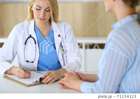 Woman doctor and patient sitting and talking at medical examination in hospital or clinic. Blonde therapist cheerfully smiling. Medicine and healthcare concept Woman doctor and patient sitting and talking at medical examination in hospital or clinic. Blonde therapist cheerfully smiling. Medicine and healthcare concept 71154123