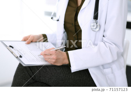 Woman doctor writing something at clipboard while sitting at the chair, close-up. Therapist at work filling up medication history records. Medicine and healthcare concept 71157123