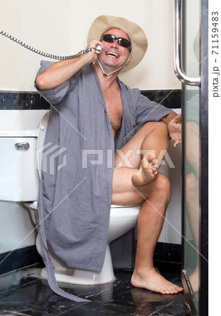 A cheerful man in the bathrobe sits on the toilet bowl and call from telephone. A cheerful man in the bathrobe sits on the toilet bowl and call from telephone. 71159483