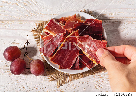 Woman hand takes a slice of jamon from a plate over white wood table.  71166535