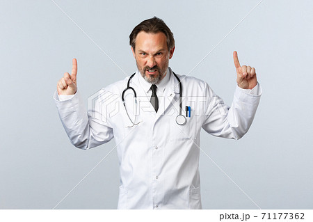 Covid-19, coronavirus outbreak, healthcare workers and pandemic concept. Angry outraged male doctor in white coat, pointing fingers up grimacing furious, looking mad at camera, white background Covid-19, coronavirus outbreak, healthcare workers and pandemic concept. Angry outraged male doctor in white coat, pointing fingers up grimacing furious, looking mad at camera, white background 71177362