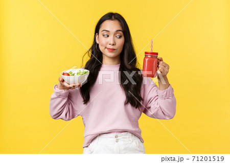 People emotions, healthy lifestyle and food concept. Curious cute asian girl looking at smoothie, holding salad and drink, making choice what eat for breakfast, standing yellow background 71201519