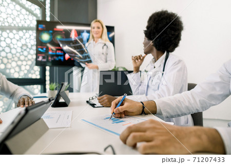 Meeting of international doctors in conference room, listening female blond speaker, holding CT scan. Focus on hands of male Indian doctor, making notes. African lady looking at lecturer Meeting of international doctors in conference room, listening female blond speaker, holding CT scan. Focus on hands of male Indian doctor, making notes. African lady looking at lecturer 71207433