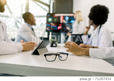 Blurred image of multiethnical concentrated medical team, having a meeting, and listening the lecture of their blond female colleague. Focus on stylish eyeglasses on the table. Education concept 71207434