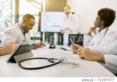 Medicine, technology concept. Blurred image of multiethnical male and female doctors having morning meeting in modern sunny office clinic. Focus on the stethoscope and digital tablet on the table 71207477