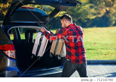 Young man unloads luggage out of the SUV after coming back home from summer family vacation. Young man unloads luggage out of the SUV after coming back home from summer family vacation. 71210790