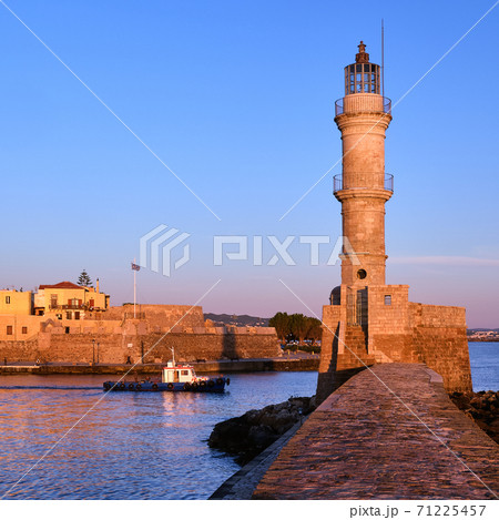 Fishing boat pass by Lighthouse lit by first sunrays in Old Venetian harbour, Chania, Crete, Greece at sunrise. Square ratio, soft early morning. 71225457