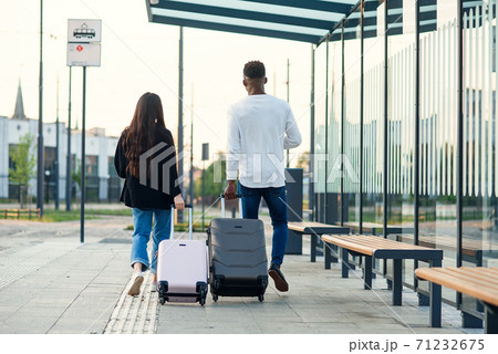 Stylish girl and black guy carrying their suitcases on wheels holding passports with tickets and walking on bus station. Stylish girl and black guy carrying their suitcases on wheels holding passports with tickets and walking on bus station. 71232675