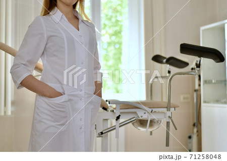 Closeup of a doctor in a white uniform, with folded hands in front of him. Stands in front of a gynecological chair. 71258048