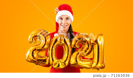 New Year 2021 Gold Balloons. Young woman in Santa Claus hat having smiling holding in hand 2021. Girl in red hat and red Dress stands on bright orange background. Concept Christmas celebration 71263036