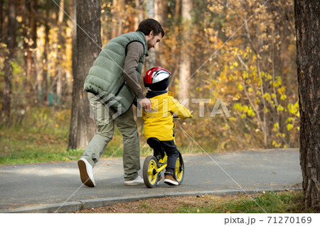 Rear view of young man running after his active little son riding balance bike Rear view of young man running after his active little son riding balance bike 71270169