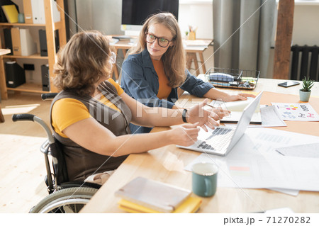 Two young female office managers sitting by table and discussing working points Two young female office managers sitting by table and discussing working points 71270282