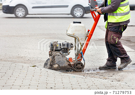 A worker cuts the old asphalt with a gas saw on the carriageway against the backdrop of a city street. 71279753