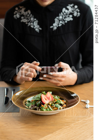 A girl in a black blouse photographs food in a restaurant with a smartphone. Vegetable salad garnished with fresh strawberries. Shallow depth of field, blurred background. A girl in a black blouse photographs food in a restaurant with a smartphone. Vegetable salad garnished with fresh strawberries. Shallow depth of field, blurred background. 71284437