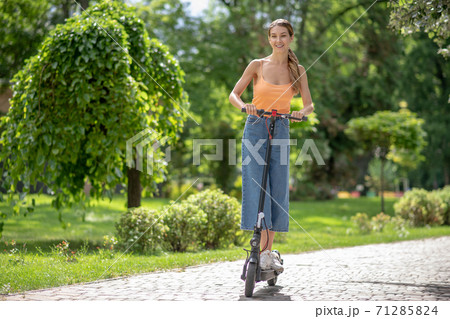 Long-haired young girl riding a scooter in the park and looking contented Long-haired young girl riding a scooter in the park and looking contented 71285824