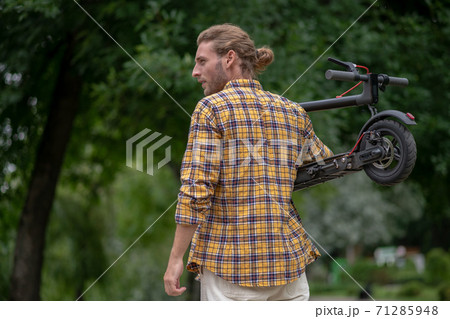 Man in checkered shirt carrying his scooter and looking aside Man in checkered shirt carrying his scooter and looking aside 71285948