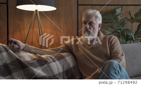 An elderly man with a gray beard sitting on the sofa turns on and off the lamp. A brooding older man is depressed 71298492
