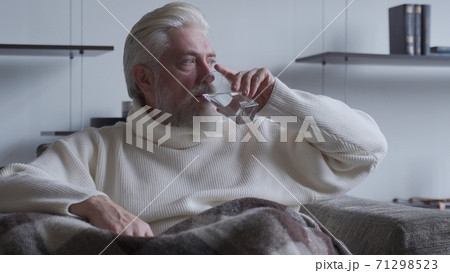 Elderly man with a gray beard is sitting in a chair and drinking a glass of water Elderly man with a gray beard is sitting in a chair and drinking a glass of water 71298523
