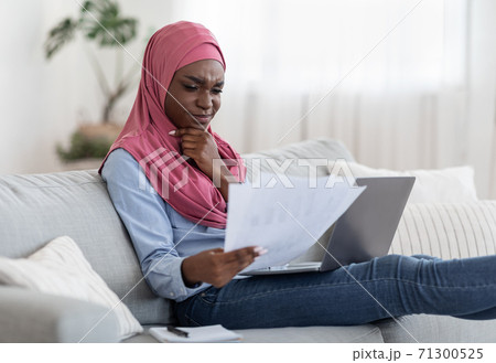 Thoughtful black muslim woman checking papers and working on laptop at home Thoughtful black muslim woman checking papers and working on laptop at home 71300525