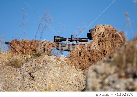 sniper in ghillie (camouflage) suit with a high-precision rifle with optic scope in a combat position in the mountains 71302870
