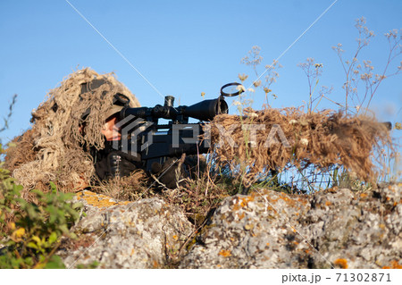 sniper in ghillie (camouflage) suit with a high-precision rifle with optic scope in a combat position in the mountains 71302871