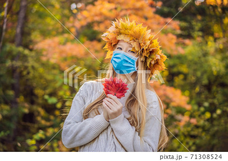 Outdoors lifestyle close up portrait of charming blonde young woman wearing a medical mask during COVID-19 coronavirus wearing a wreath of autumn leaves. Smiling, walking on the autumn park. Wearing Outdoors lifestyle close up portrait of charming blonde young woman wearing a medical mask during COVID-19 coronavirus wearing a wreath of autumn leaves. Smiling, walking on the autumn park. Wearing 71308524
