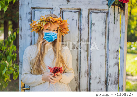 Outdoors lifestyle close up portrait of charming blonde young woman wearing a medical mask during COVID-19 coronavirus wearing a wreath of autumn leaves. Wearing stylish knitted pullover. Wreath of Outdoors lifestyle close up portrait of charming blonde young woman wearing a medical mask during COVID-19 coronavirus wearing a wreath of autumn leaves. Wearing stylish knitted pullover. Wreath of 71308527