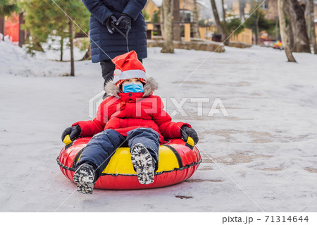mom son wearing a medical mask during COVID-19 coronavirus ride on an inflatable winter sled tubing. Winter fun for the whole family 71314644