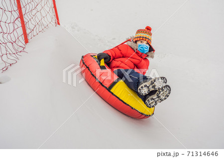 Child wearing a medical mask during COVID-19 coronavirus having fun on snow tube. Boy is riding a tubing. Winter fun for children Child wearing a medical mask during COVID-19 coronavirus having fun on snow tube. Boy is riding a tubing. Winter fun for children 71314646