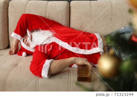 Drunk santa claus is sleeping on couch with bottle of whiskey in hands at home Drunk santa claus is sleeping on couch with bottle of whiskey in hands at home 71323244