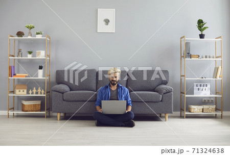 Young smiling man sitting on floor with laptop on knees and working online from home Young smiling man sitting on floor with laptop on knees and working online from home 71324638