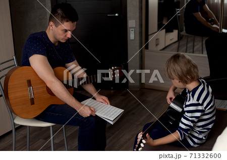 Young male musician teaches little boy student how to play the acoustic guitar and play by notes Young male musician teaches little boy student how to play the acoustic guitar and play by notes 71332600