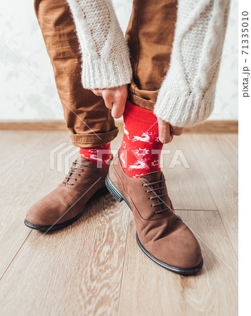 Young man pulls up leg of his chinos trousers to show bright red socks with reindeers on them. Scandinavian pattern. Winter holiday spirit. Casual outfit for New Year and Christmas celebration. Young man pulls up leg of his chinos trousers to show bright red socks with reindeers on them. Scandinavian pattern. Winter holiday spirit. Casual outfit for New Year and Christmas celebration. 71335010